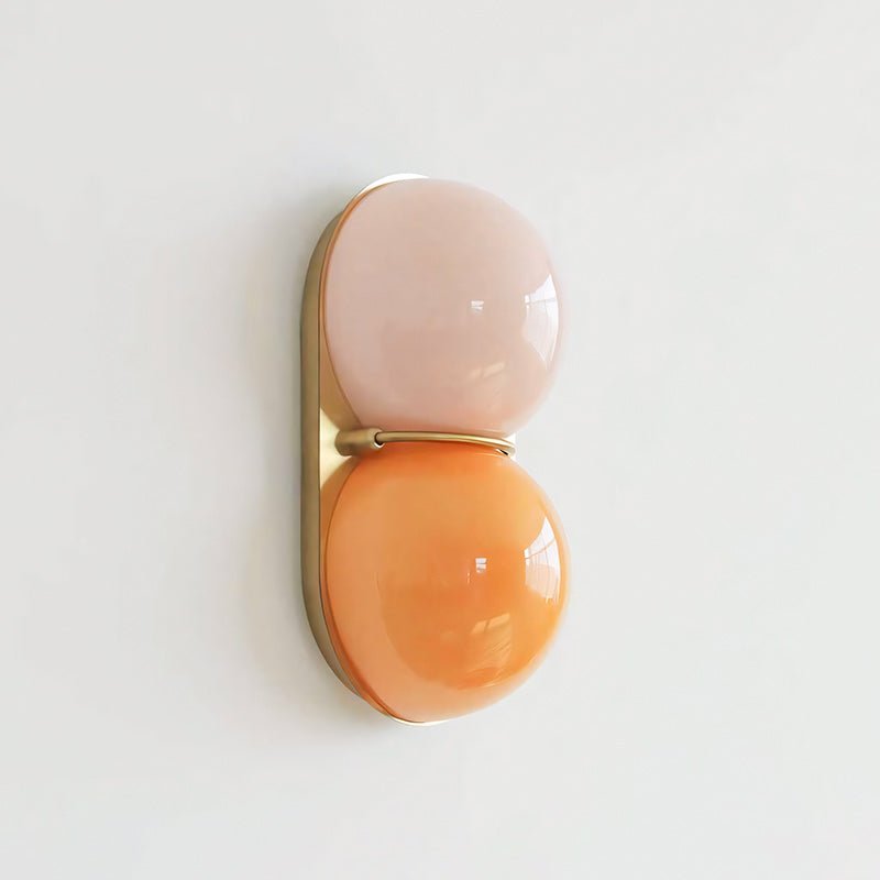Blush-pink globe above an orange globe, this Clérachic wall light has a gold-toned backplate and sits on a pale wall.