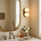 Beside an oval gold-framed mirror, a black-backed Clérachic sconce glows warm above a marble vanity with brass taps.