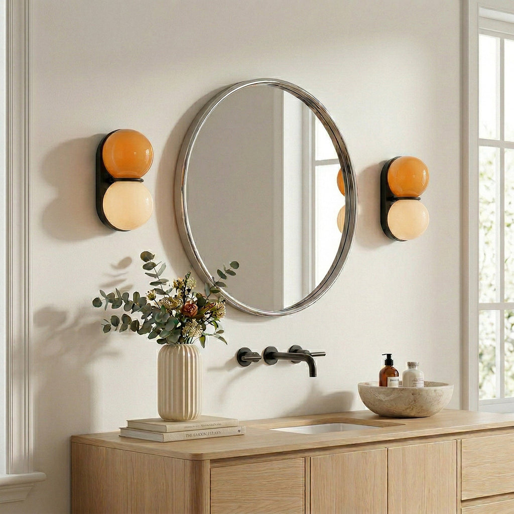 A pair of Clérachic wall lights with orange upper globes flank a round mirror above a pale wood vanity with a wall-mounted tap.
