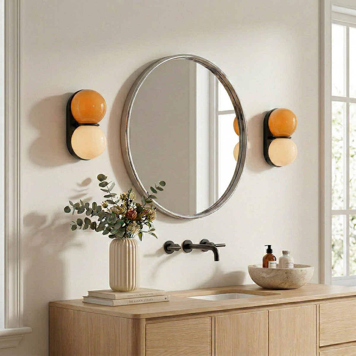A pair of Clérachic wall lights with orange upper globes flank a round mirror above a pale wood vanity with a wall-mounted tap.
