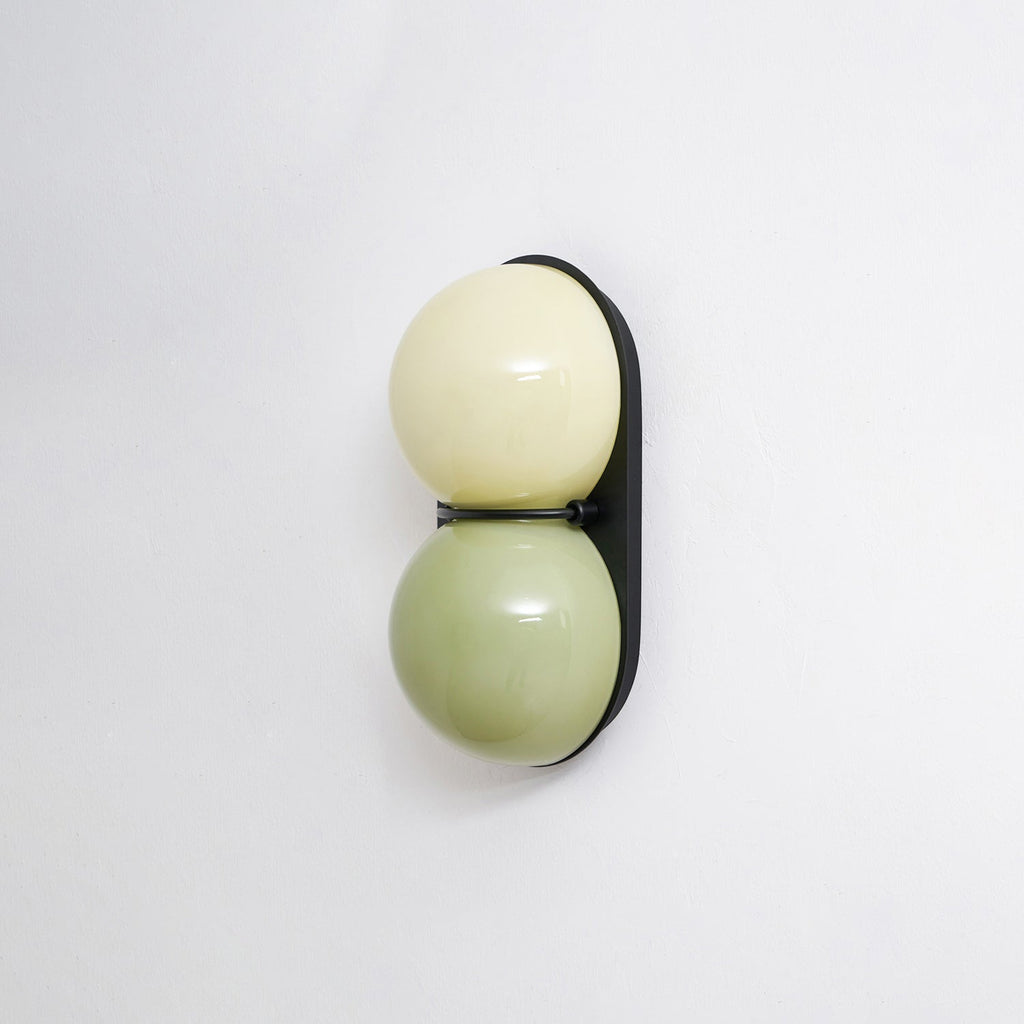 Sage green below pale cream, these glass globes sit on a black oval backplate on this Clérachic wall light, shot against white.