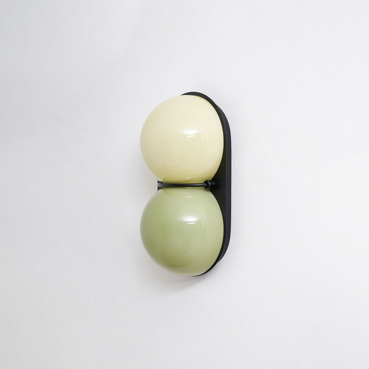 Sage green below pale cream, these glass globes sit on a black oval backplate on this Clérachic wall light, shot against white.