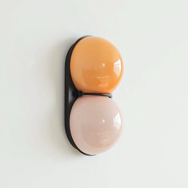 Apricot upper globe above a pale blush one on a black oval plate, this Clérachic sconce is seen in profile on a pale wall.