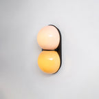 Lit warm, this Clérachic sconce has a pale pink upper globe and a golden-yellow lower globe on a black backplate.