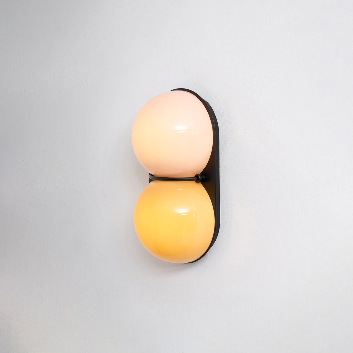 Lit warm, this Clérachic sconce has a pale pink upper globe and a golden-yellow lower globe on a black backplate.