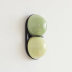 Sage-green over pale lime on a black backplate, this Clérachic wall light hangs on a white wall in soft daylight.