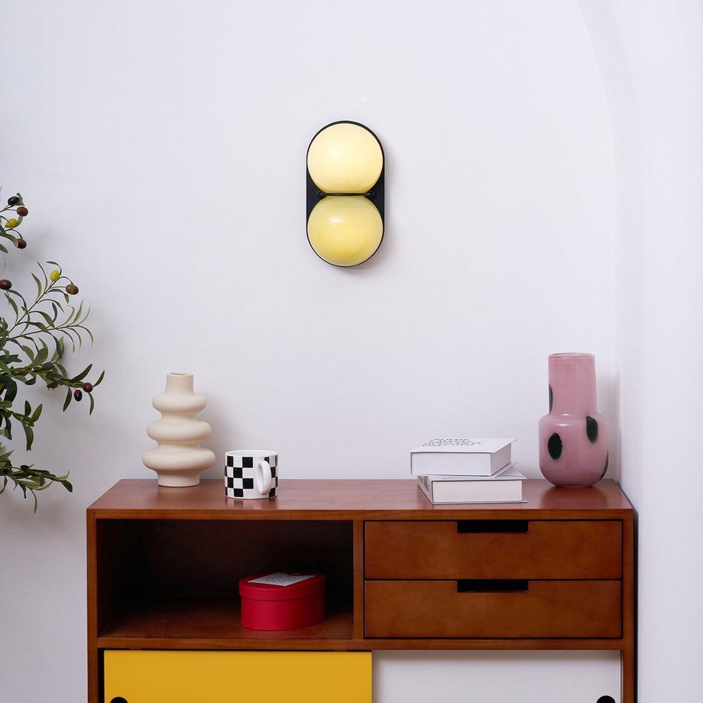 High on a white wall above a walnut sideboard, this Clérachic sconce has pale yellow glass globes on a black plate.