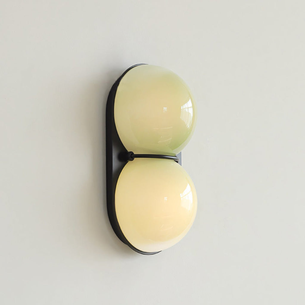 Slightly turned view of a Clérachic sconce with two soft yellow-green globes on a black plate against a pale grey wall.