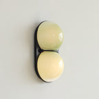 Slightly turned view of a Clérachic sconce with two soft yellow-green globes on a black plate against a pale grey wall.