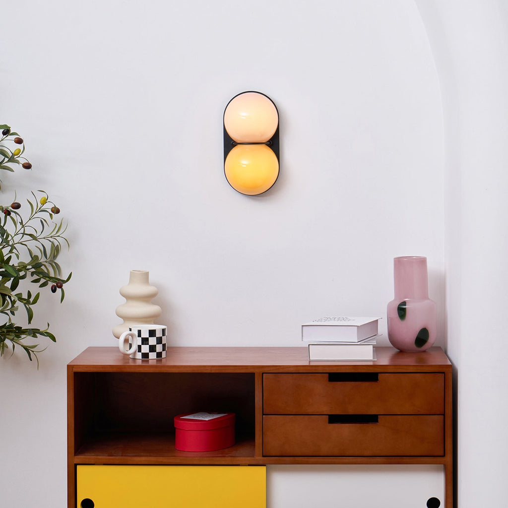 Glowing peach and amber globes on a Clérachic wall light high above a walnut sideboard with books and a checked mug.