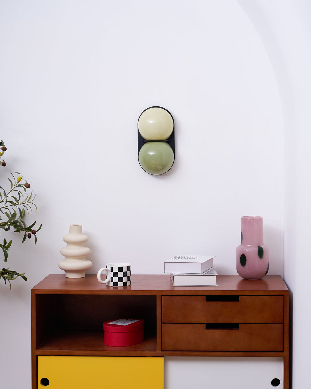 Small in the frame, a sage and cream Clérachic wall light hangs above a walnut sideboard with a red box and pink vase.