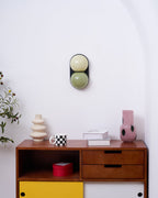 Small in the frame, a sage and cream Clérachic wall light hangs above a walnut sideboard with a red box and pink vase.