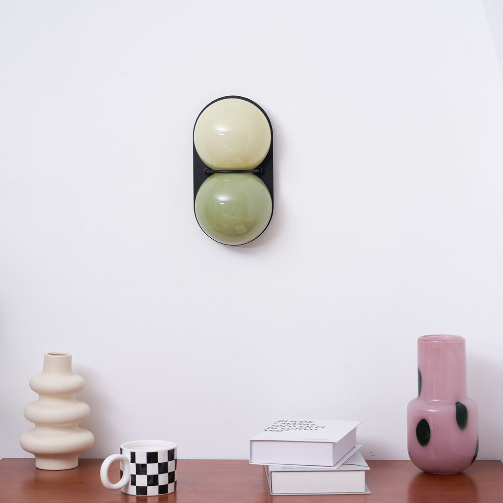 Pale cream upper globe over sage green, this Clérachic wall light is shown close up above a walnut sideboard and cream vase.