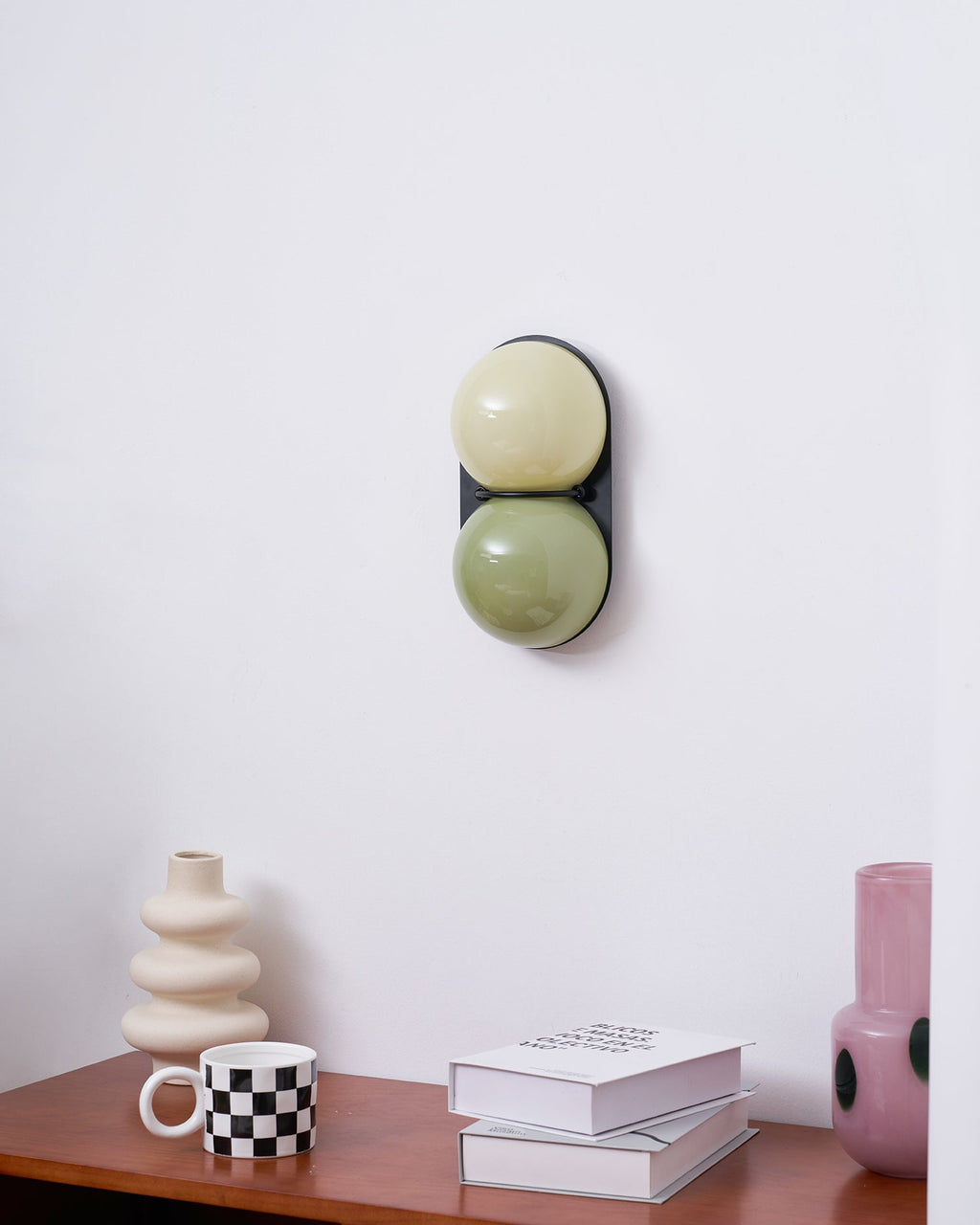 A sage-and-cream Clérachic sconce on a black plate, seen from the side with a ribbed cream vase and a pink spotted vase below.