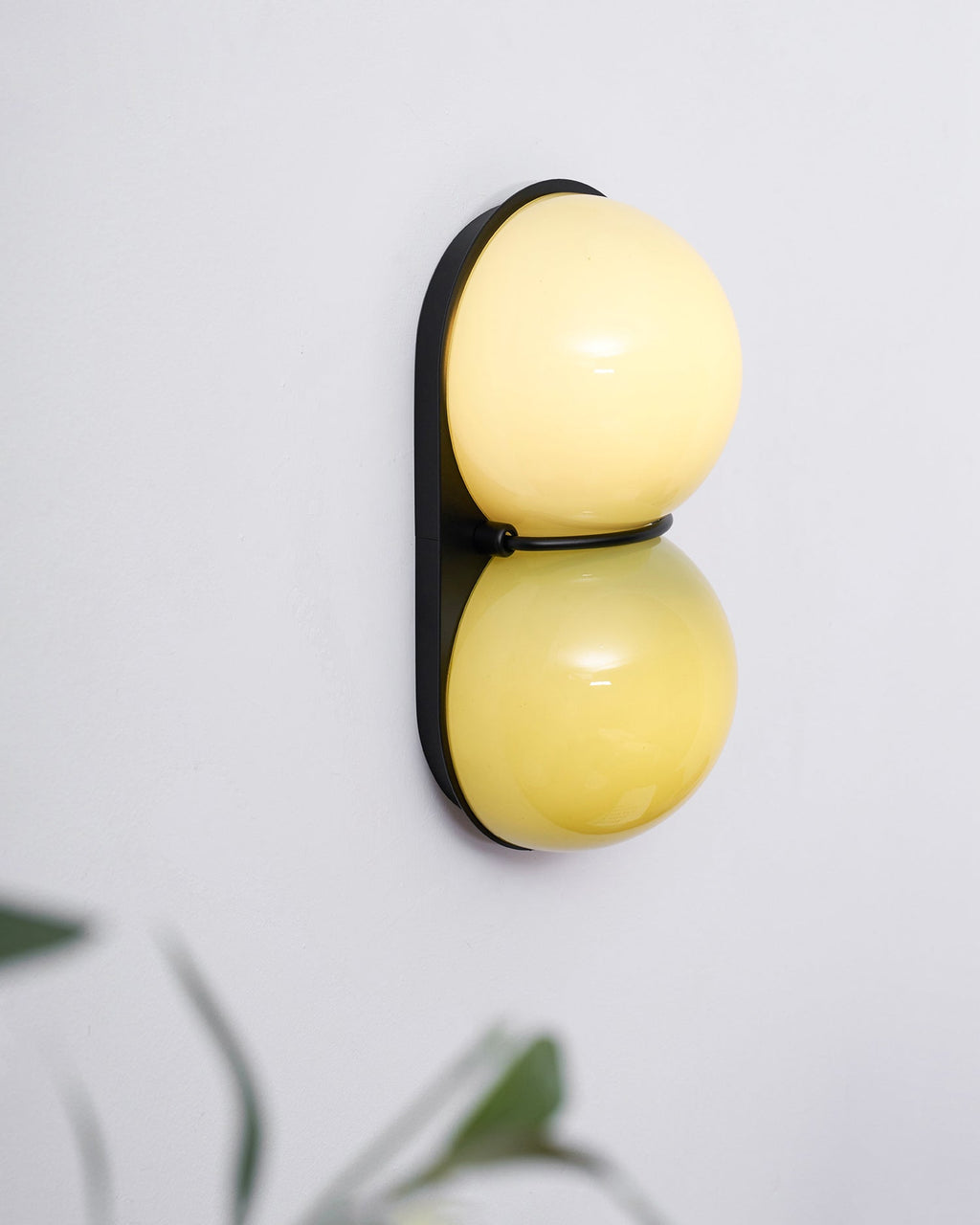 Lit Clérachic sconce with a bright cream upper globe and a golden-yellow lower globe, with soft leaves blurred in front.
