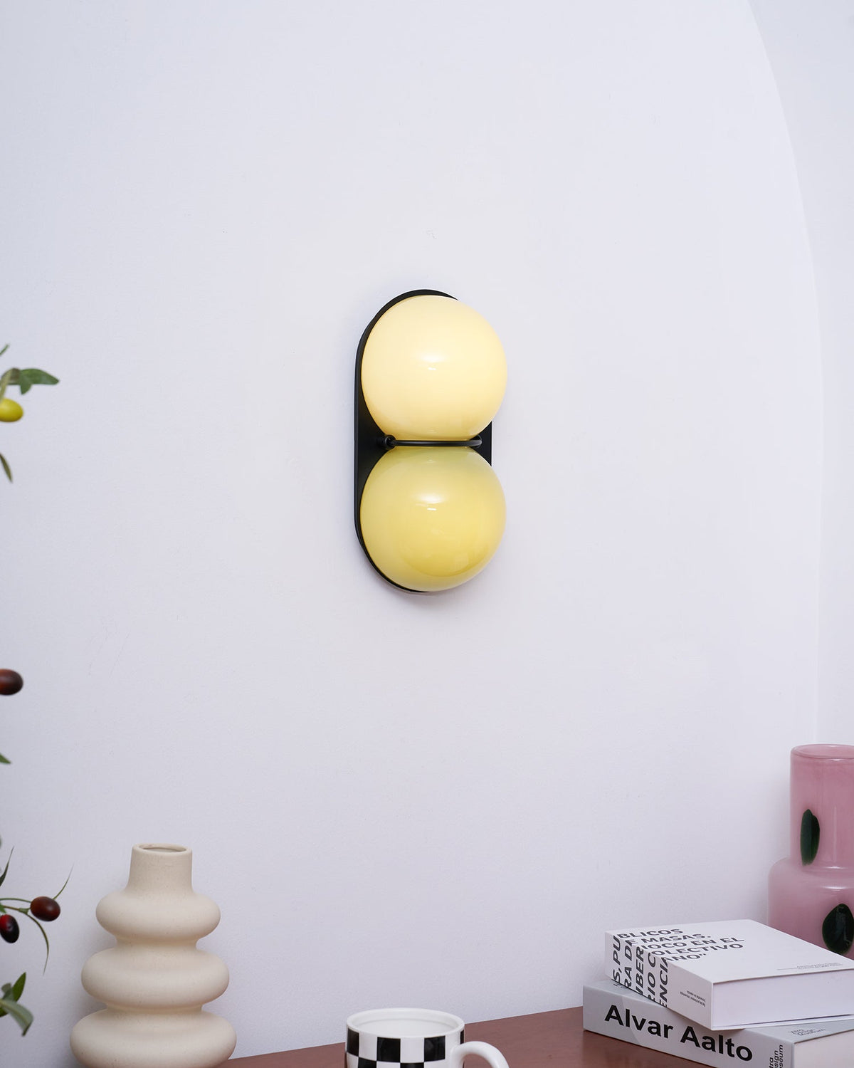 On a white wall above a desk with books, this Clérachic wall light glows cream over lemon yellow on its black backplate.