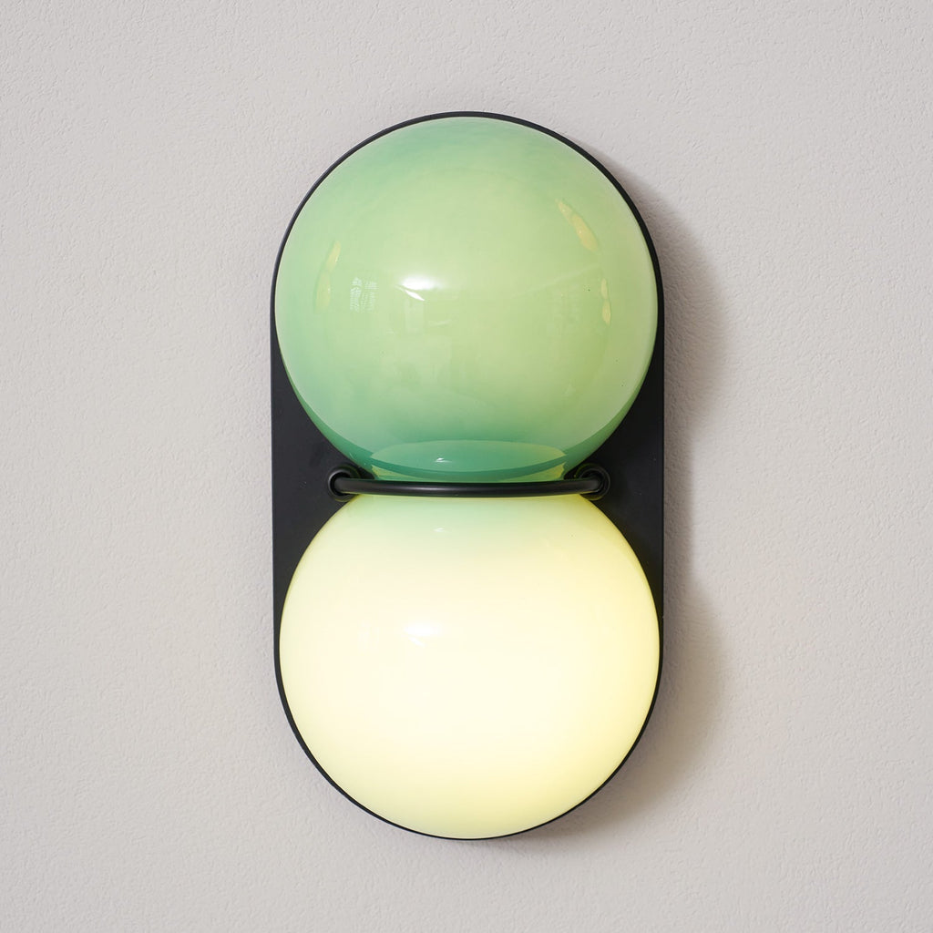 Mint-green glass over a glowing cream globe on a black oval plate, this Clérachic wall light is shown close up on a grey wall.