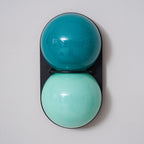 Front view of a Clérachic sconce with a deep teal globe over aqua on a black oval plate, against a pale grey wall.