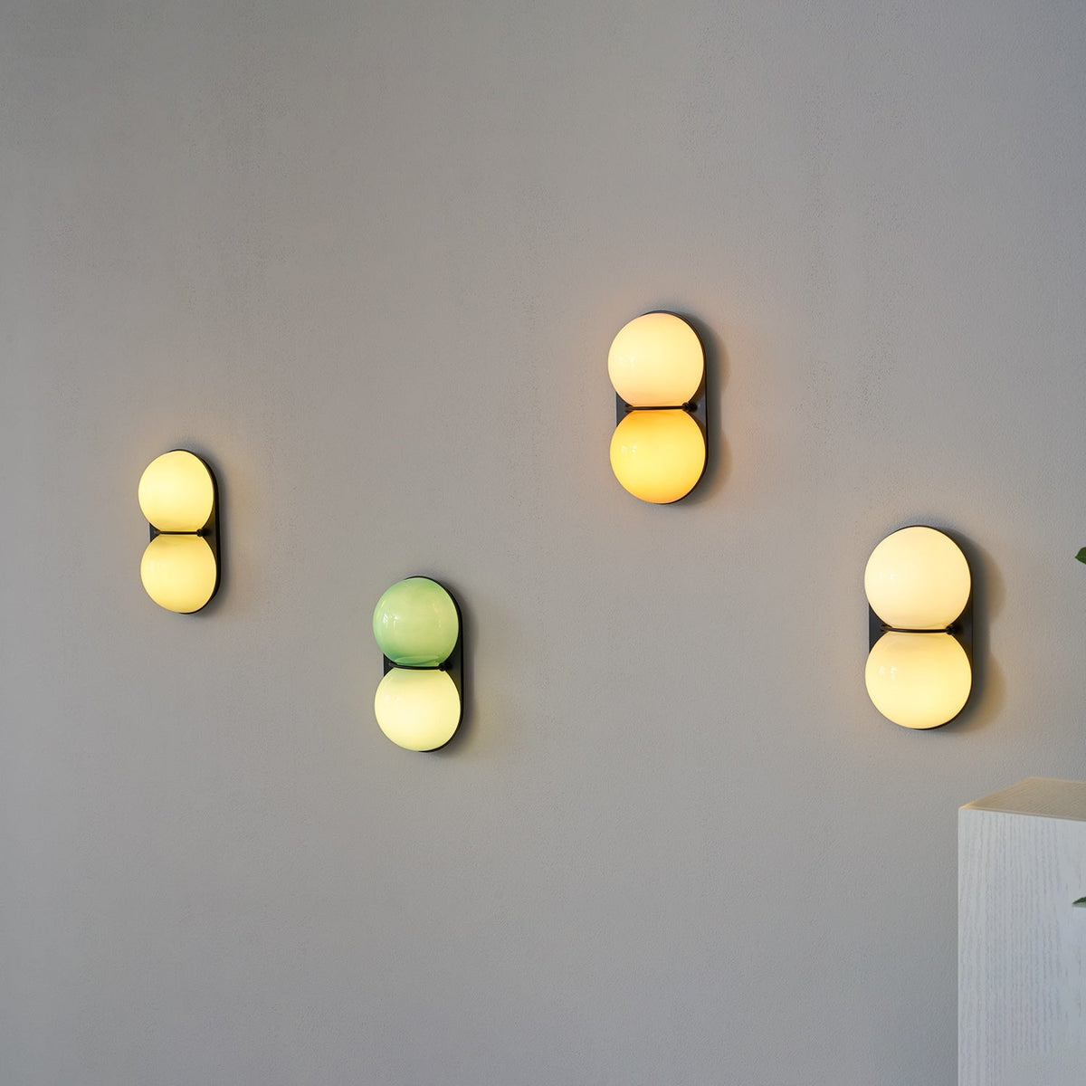 Four lit Clérachic wall lights scattered on a grey wall, one sage-green and the rest warm cream, yellow and white.