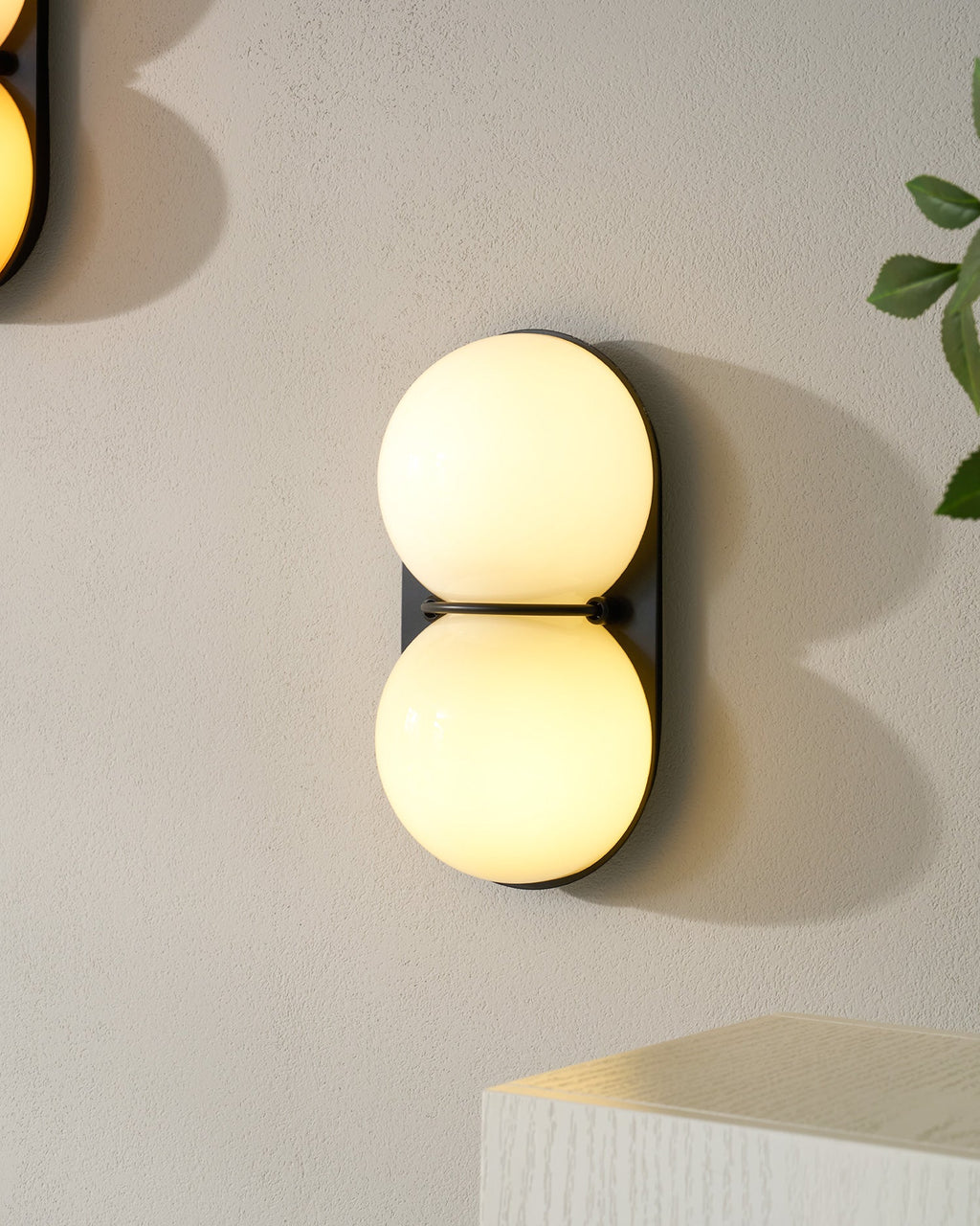 One lit cream Clérachic wall light close up on a pale wall, with another sconce edging into the corner of the frame.