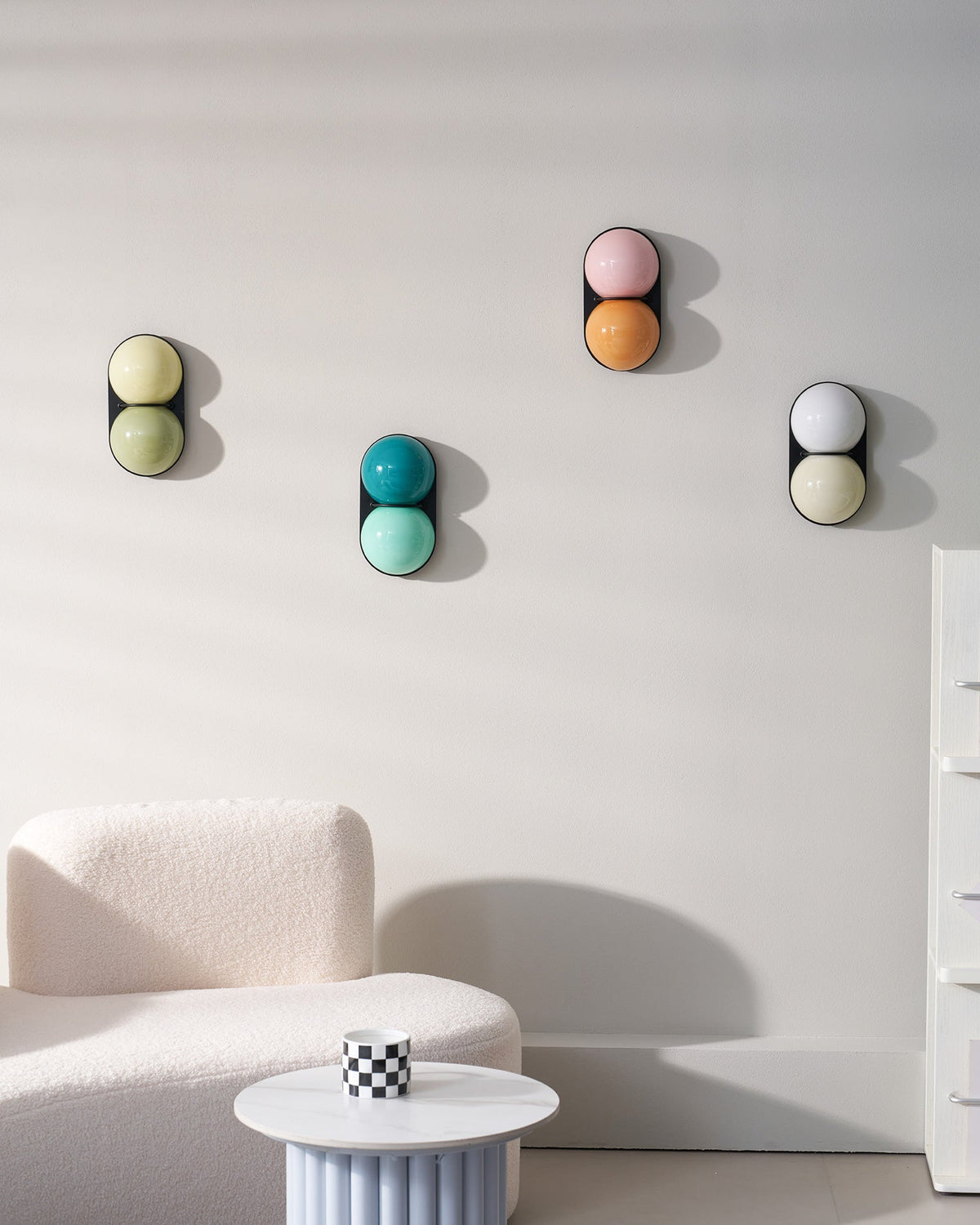 Four unlit Clérachic wall lights in sage, teal, pink-orange and white-cream arranged on a white wall above a curved sofa.