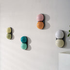 Unlit Clérachic wall lights in olive, teal, pink-orange and grey-white stagger across a white wall near a pale shelf.