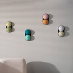 Four unlit Clérachic wall lights in different glass colours climb a pale grey wall beside a cream bouclé seat.