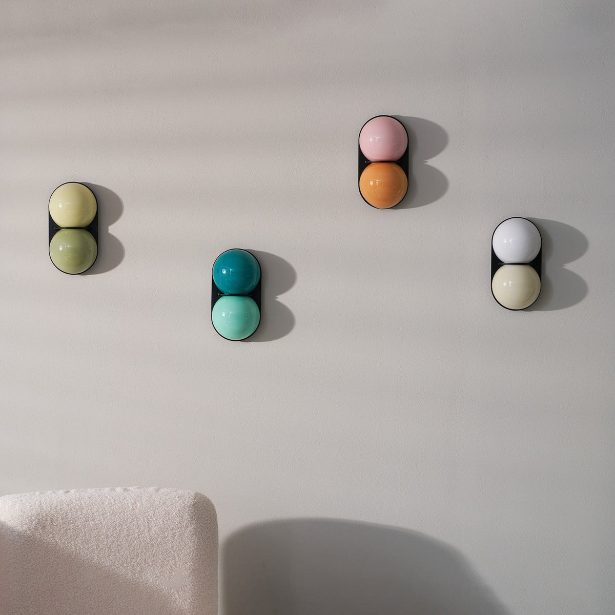 Four unlit Clérachic wall lights in different glass colours climb a pale grey wall beside a cream bouclé seat.