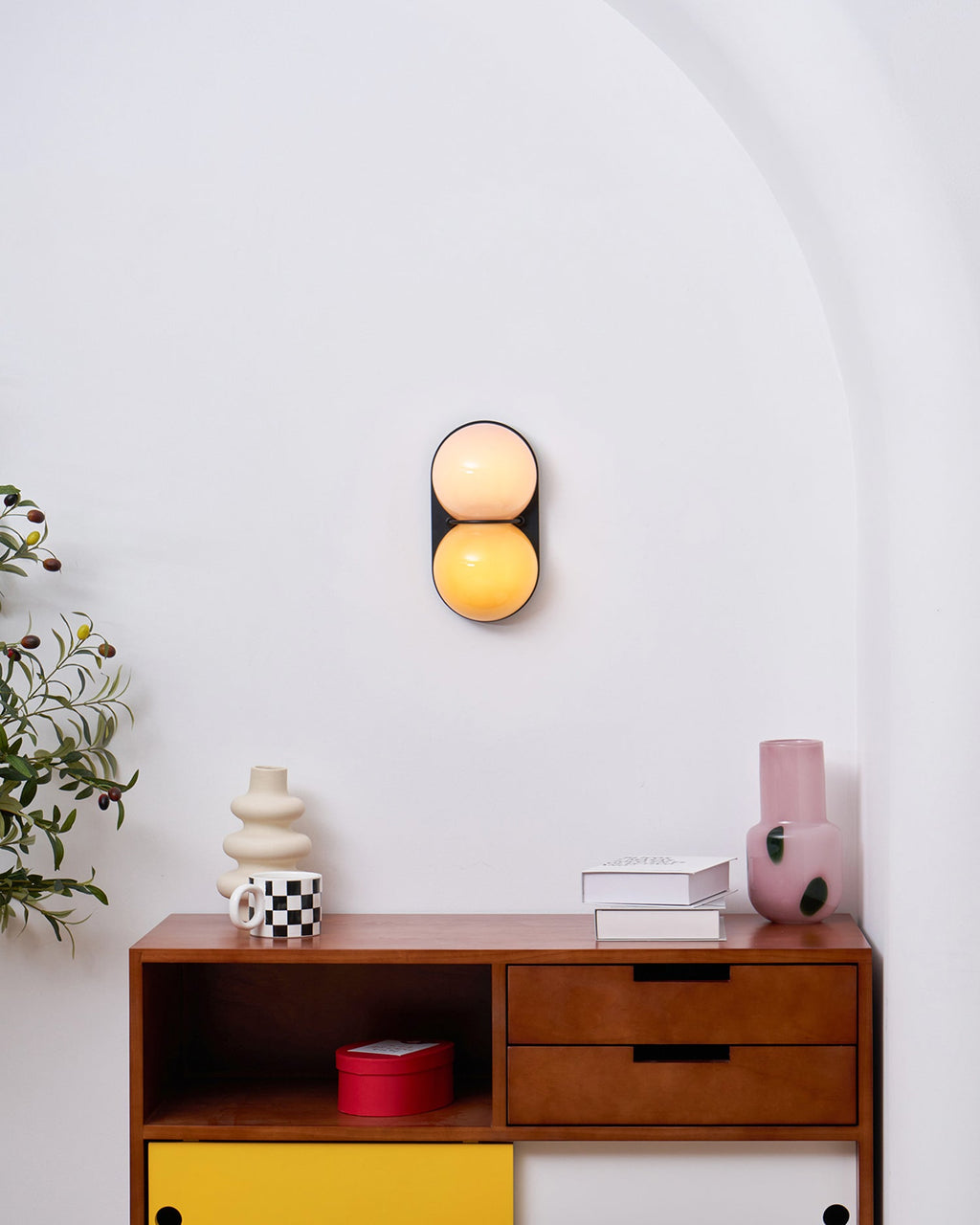 On a white wall above a walnut sideboard, a lit Clérachic sconce shows a peach globe over an amber one.