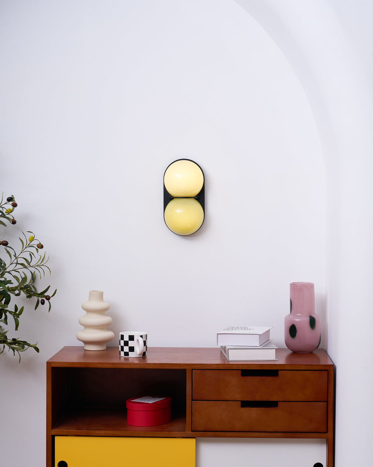 Lit Clérachic sconce in cream and lemon yellow, mounted above a walnut sideboard with a pink vase and a cream vase.