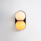 Close front view of a lit Clérachic wall light, a blush upper globe over a warm amber one on a black oval plate.