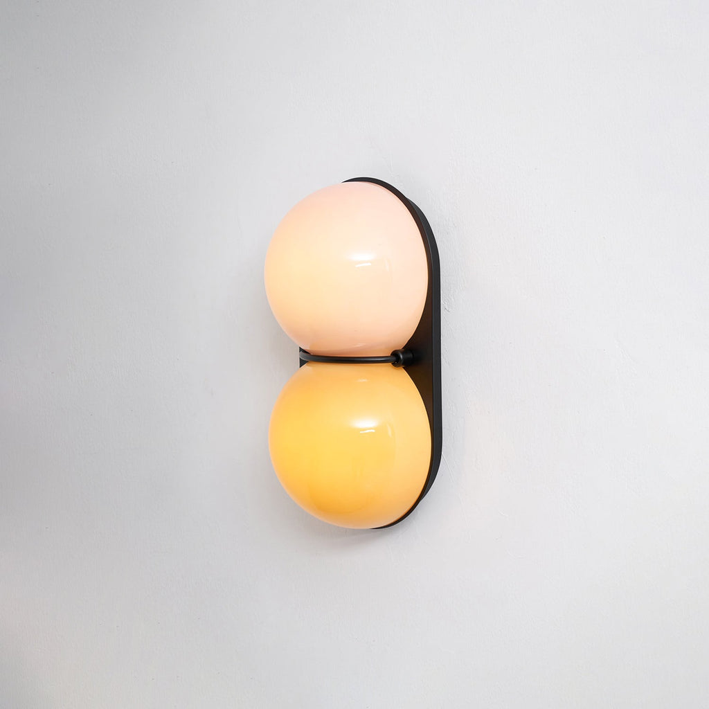 Slightly angled Clérachic sconce glowing peach above amber against a plain pale wall, its black plate casting a soft shadow.