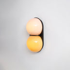 Slightly angled Clérachic sconce glowing peach above amber against a plain pale wall, its black plate casting a soft shadow.