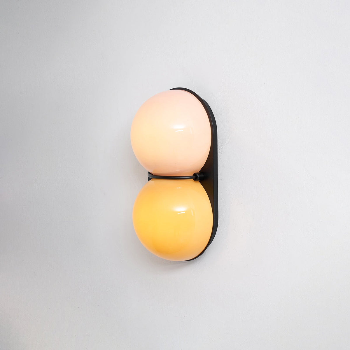 Slightly angled Clérachic sconce glowing peach above amber against a plain pale wall, its black plate casting a soft shadow.