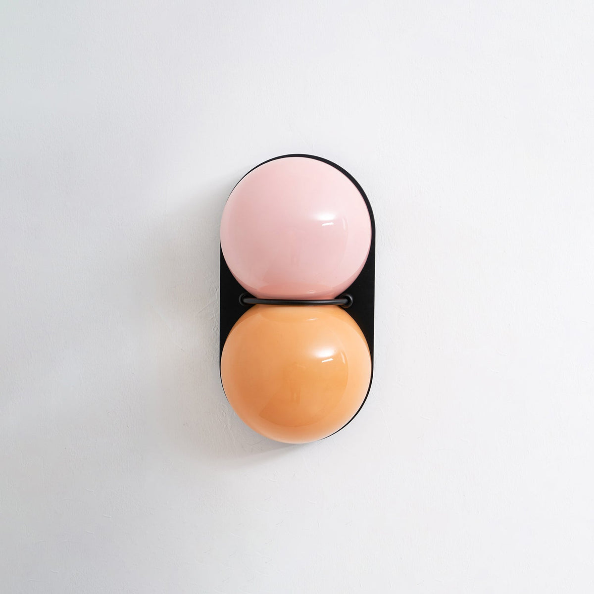 A pink upper globe over a soft orange one on a black oval plate, this Clérachic wall light unlit on a pale grey wall.