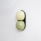 Three-quarter angle on a Clérachic sconce with pale cream over sage green, on a black plate with a soft shadow behind.