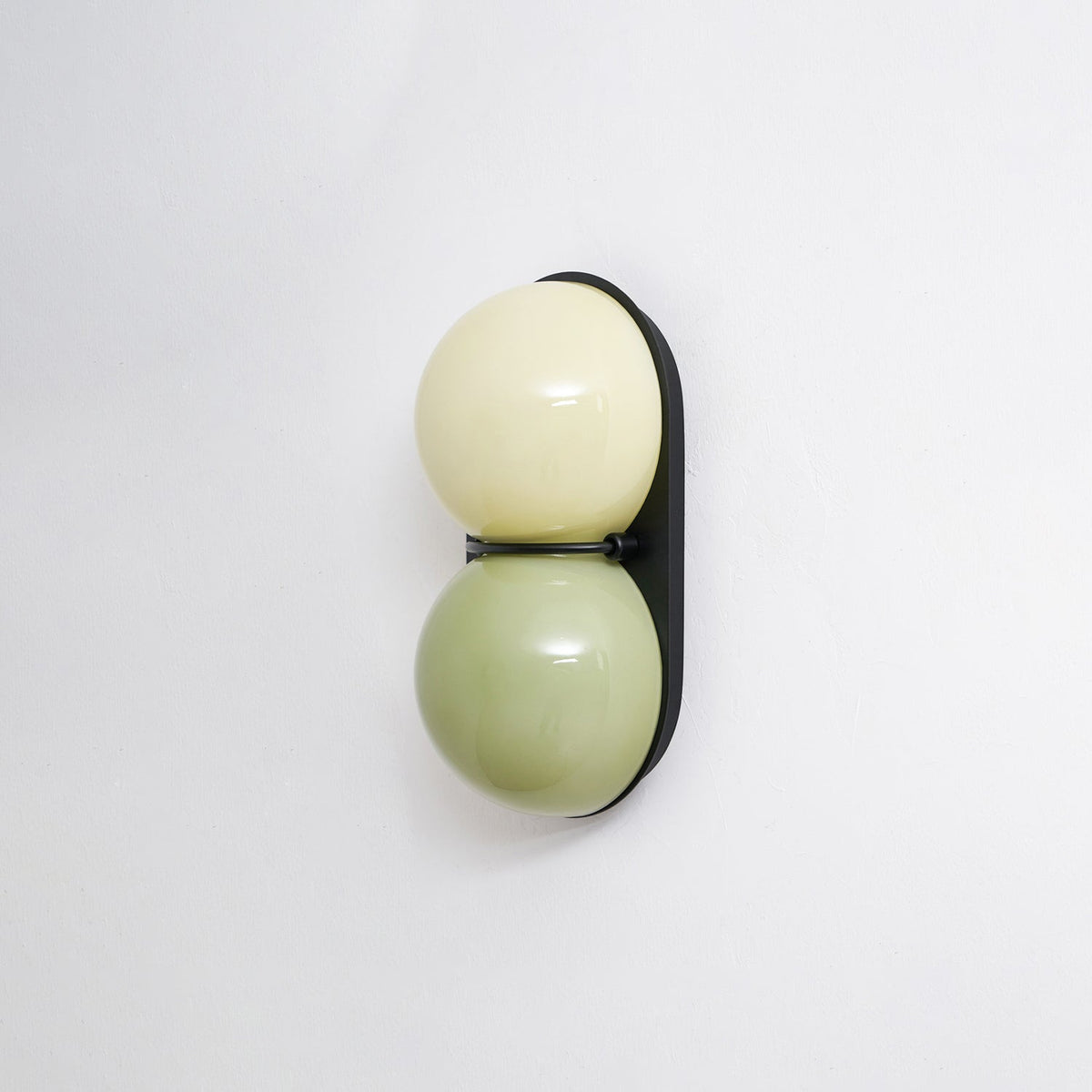 Three-quarter angle on a Clérachic sconce with pale cream over sage green, on a black plate with a soft shadow behind.