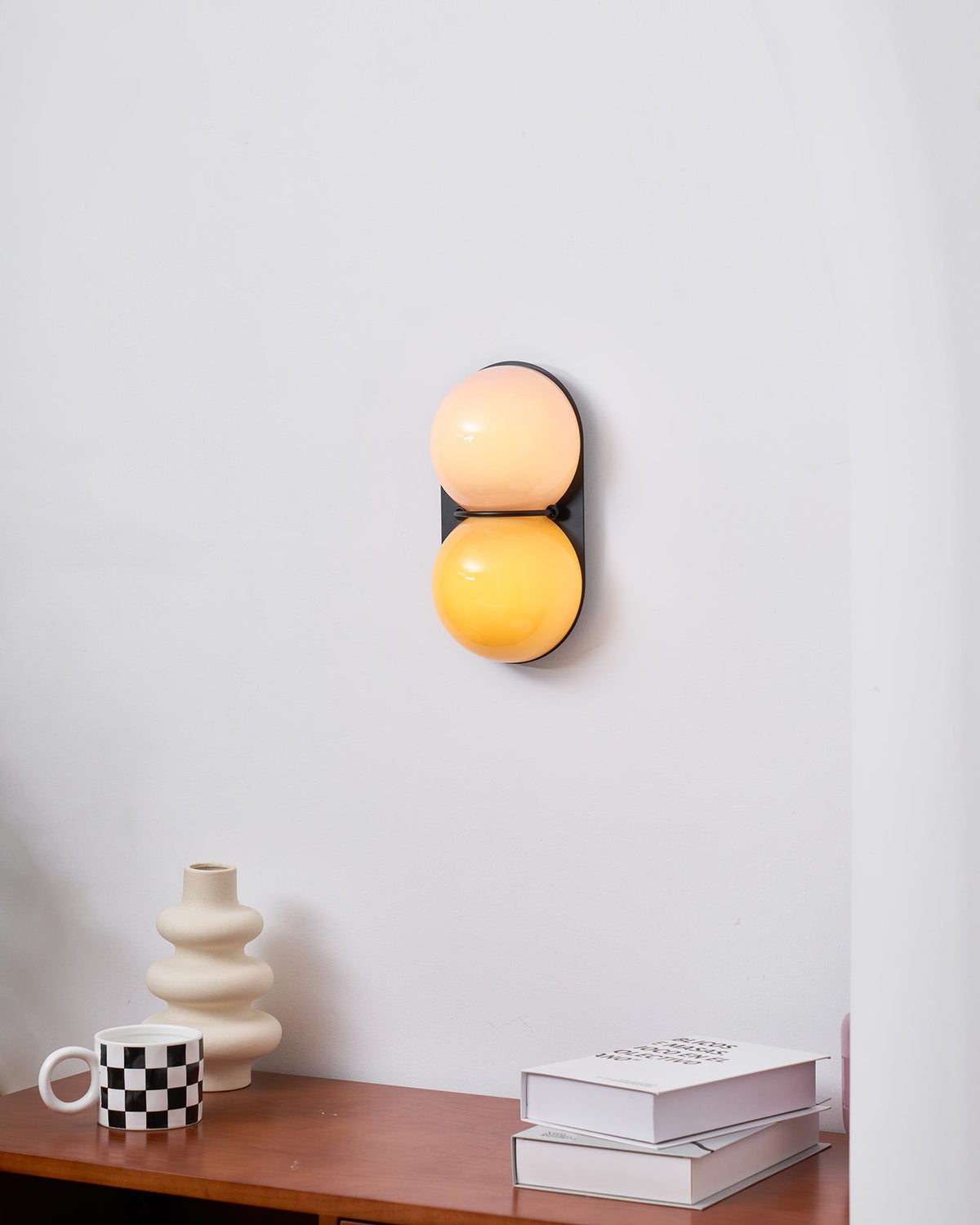 Hung on a white wall over a walnut desk, a Clérachic sconce glows peach and amber beside a cream vase and a checked cup.