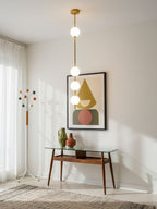 A Clérachic brass-column pendant with four globes hanging beside a framed geometric print above a glass-topped console table.