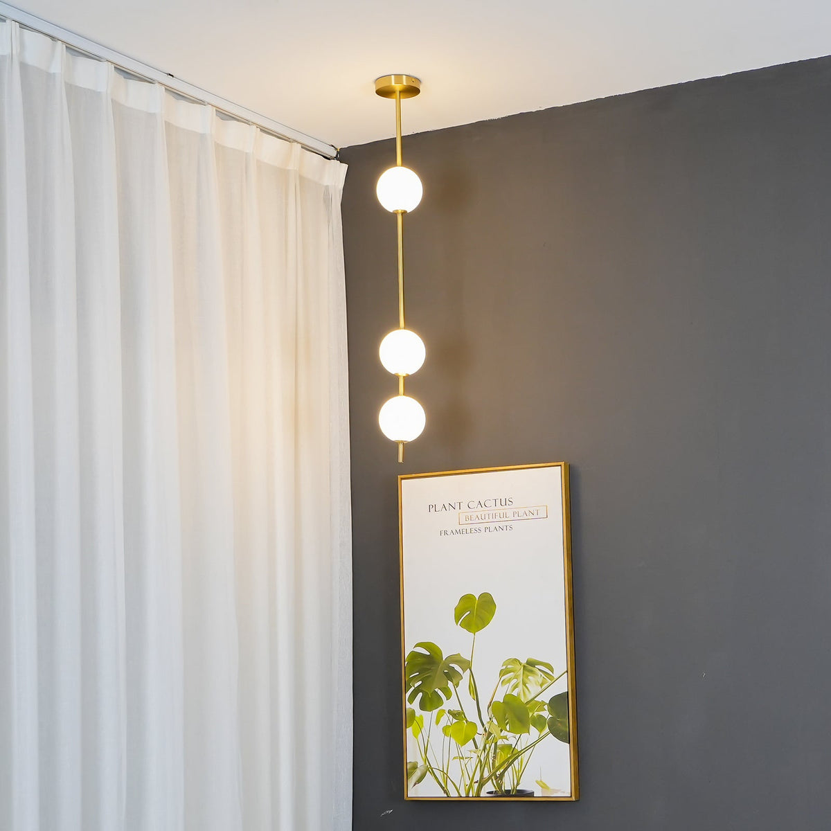 Three-globe Clérachic pendant in front of a dark grey wall, with a pale curtain on one side and a framed plant print leaning below.