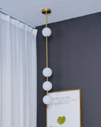 Close view of a brass-column Clérachic pendant with four opal globes, against a grey wall beside a white curtain and a framed plant print.