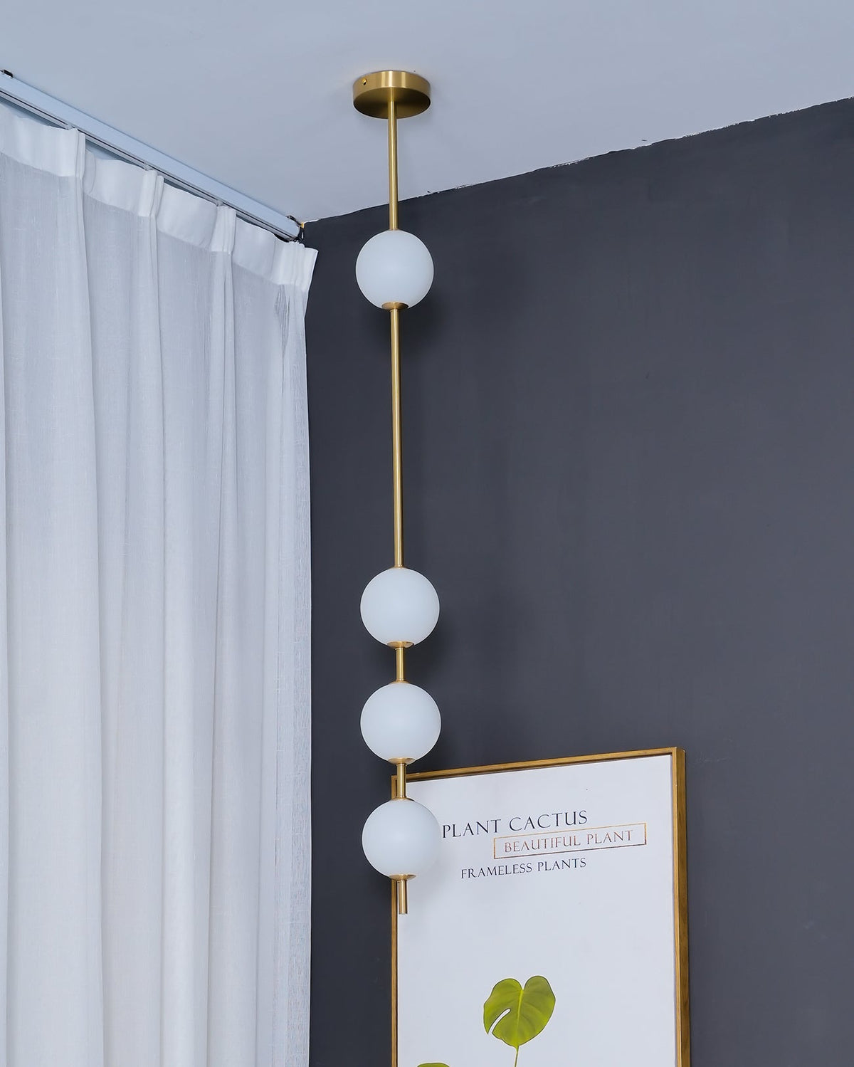 Close view of a brass-column Clérachic pendant with four opal globes, against a grey wall beside a white curtain and a framed plant print.