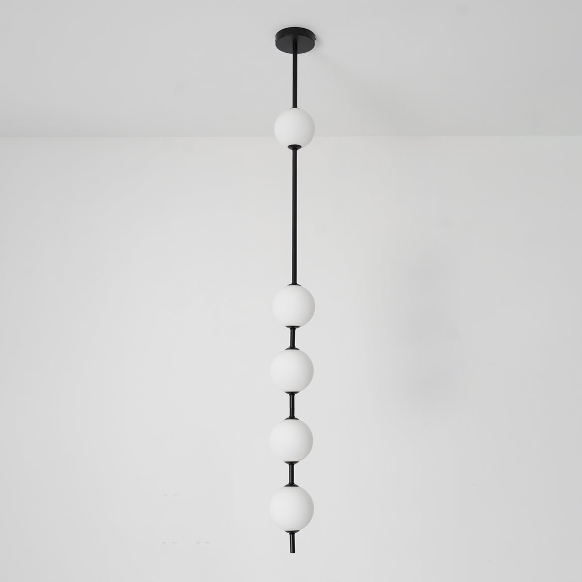 Black-column Clérachic pendant with five opal globes, the top one near a black ceiling plate, in front of a plain white wall.