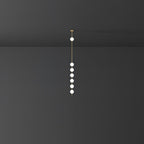 A tall, slender Clérachic pendant with six lit globes on a brass rod, shown far off against a dark grey wall.