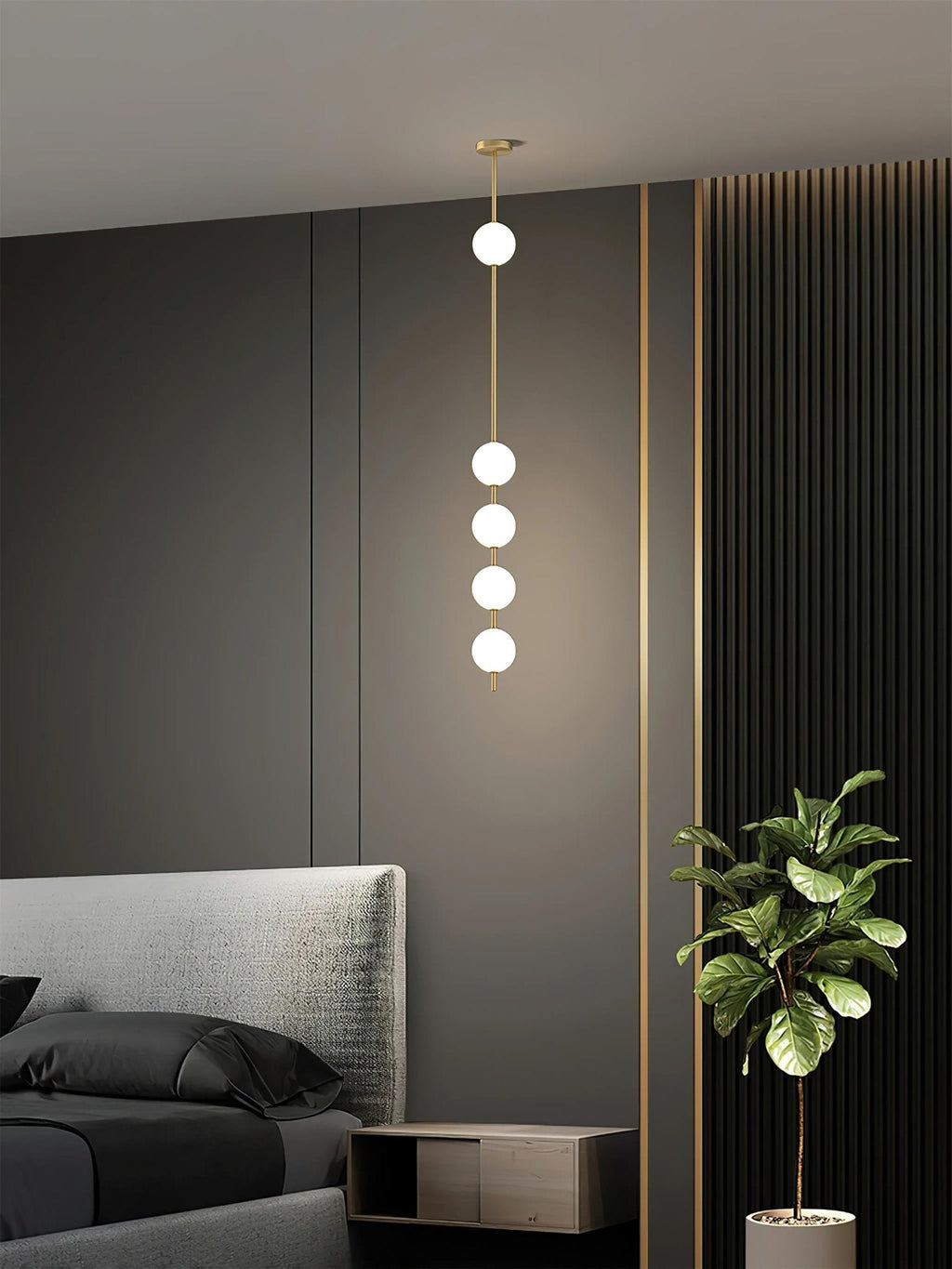 Four-globe Clérachic pendant with a brass column hung beside a bed with a grey headboard, near a dark wall and a leafy plant.