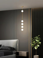 Four-globe Clérachic pendant with a brass column hung beside a bed with a grey headboard, near a dark wall and a leafy plant.