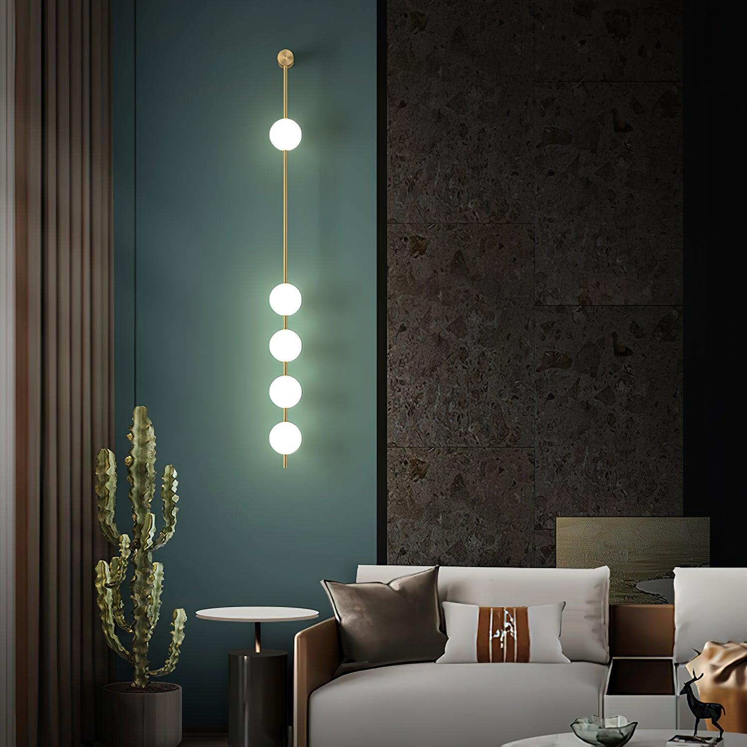 The Clérachic Stacked Globe Wall Sconce in brass, lit, five globes on a teal wall beside a cactus and side table.