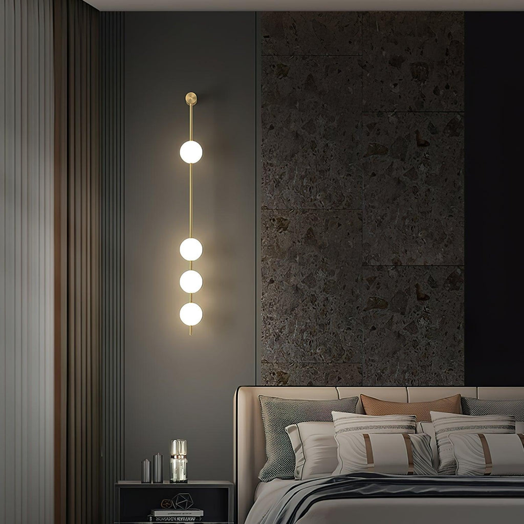 The Clérachic Stacked Globe Wall Sconce in brass, lit, four globes beside a bed against a dark stone feature wall.