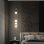 The Clérachic Stacked Globe Wall Sconce in brass, lit, four globes beside a bed against a dark stone feature wall.