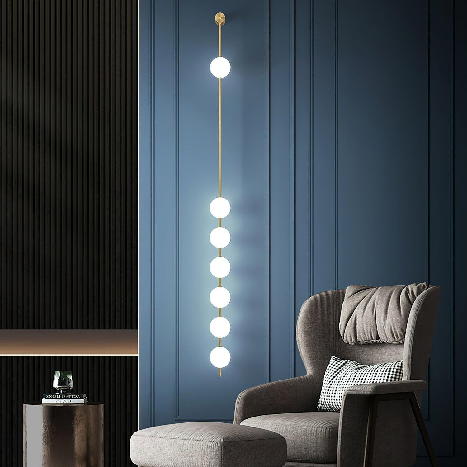 The Clérachic Stacked Globe Wall Sconce in brass, lit, six globes on a navy panelled wall beside a grey armchair.